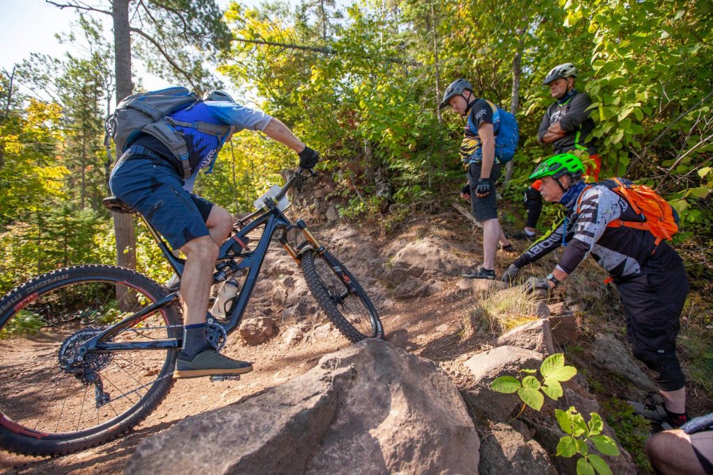 Men in bike helmets watch as a man on a mountain bike rides a bermed bank on a trail