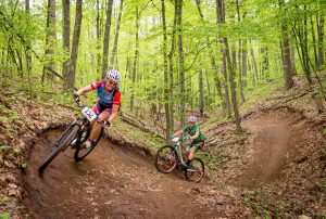 A woman rides a mountain bike on a banked turn through the forest with a man following her on a mtb.